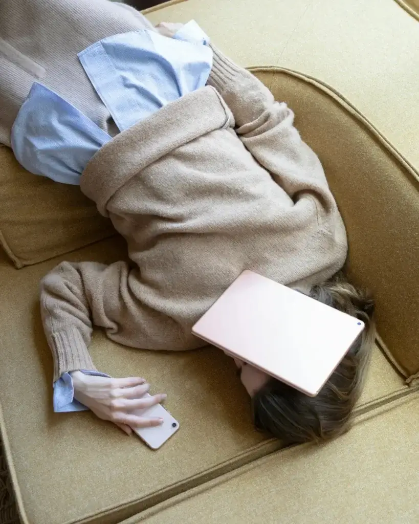Kvinde i oversized sweater ligger med hovedet nedad på en sofa med iPad på ansigtet og mobil i hånden – total digital udmattelse vibes